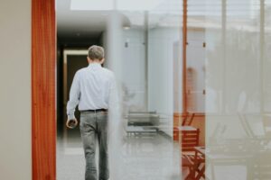 Man in a striped shirt walking away down a glass-walled office corridor