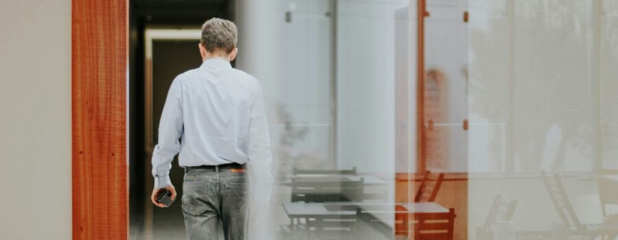 Man in a striped shirt walking away down a glass-walled office corridor